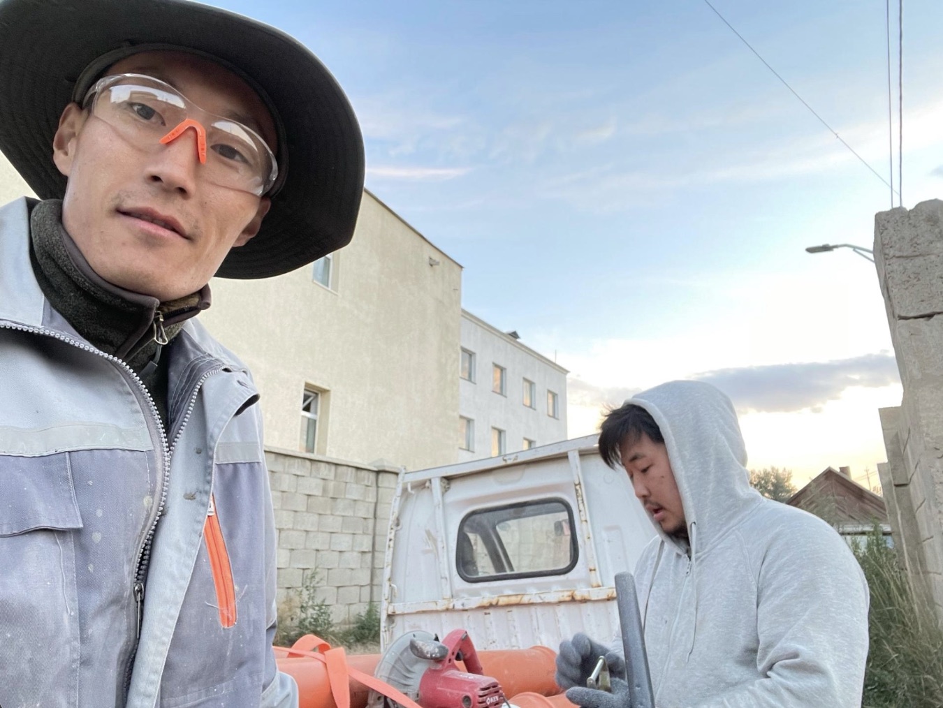 Two co-founders working outdoors beside construction equipment, one wearing work clothes and protective glasses.