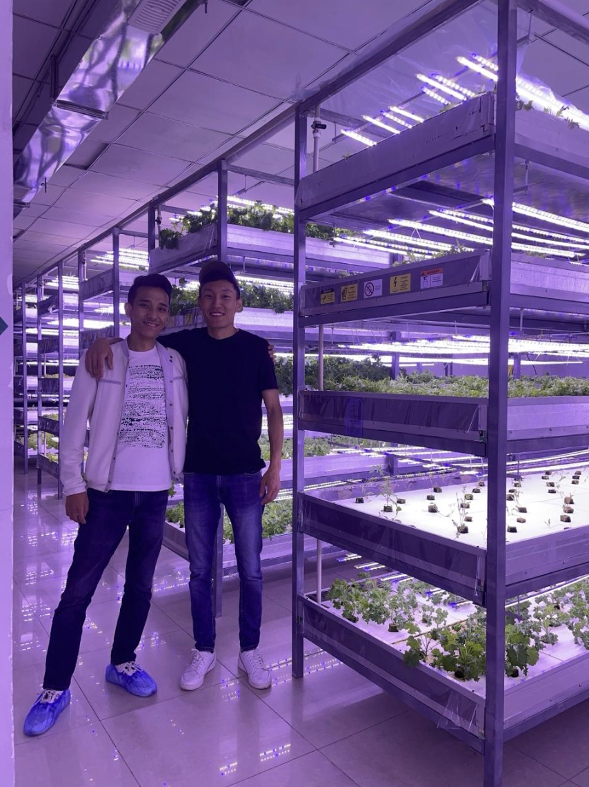 Two thin greens co-founders standing beside stacked hydroponic growing shelves under purple grow lights in their pilot farm.